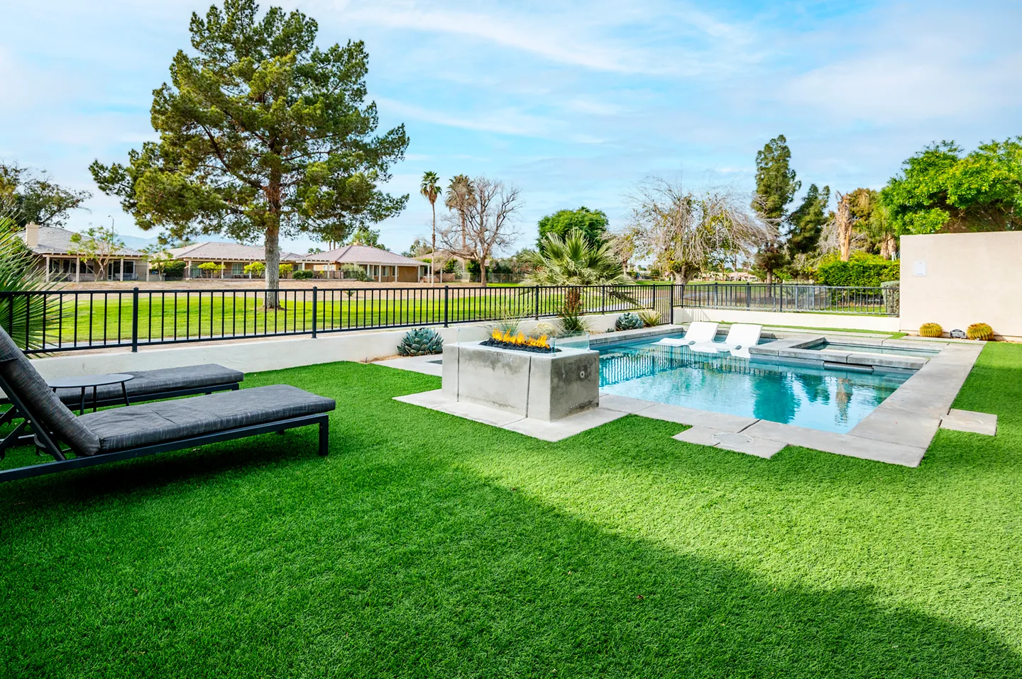 Backyard with artificial turf lawn, swimming pool with in-pool loungers, hot tub, modern fire pit, and black metal fencing overlooking a golf course.