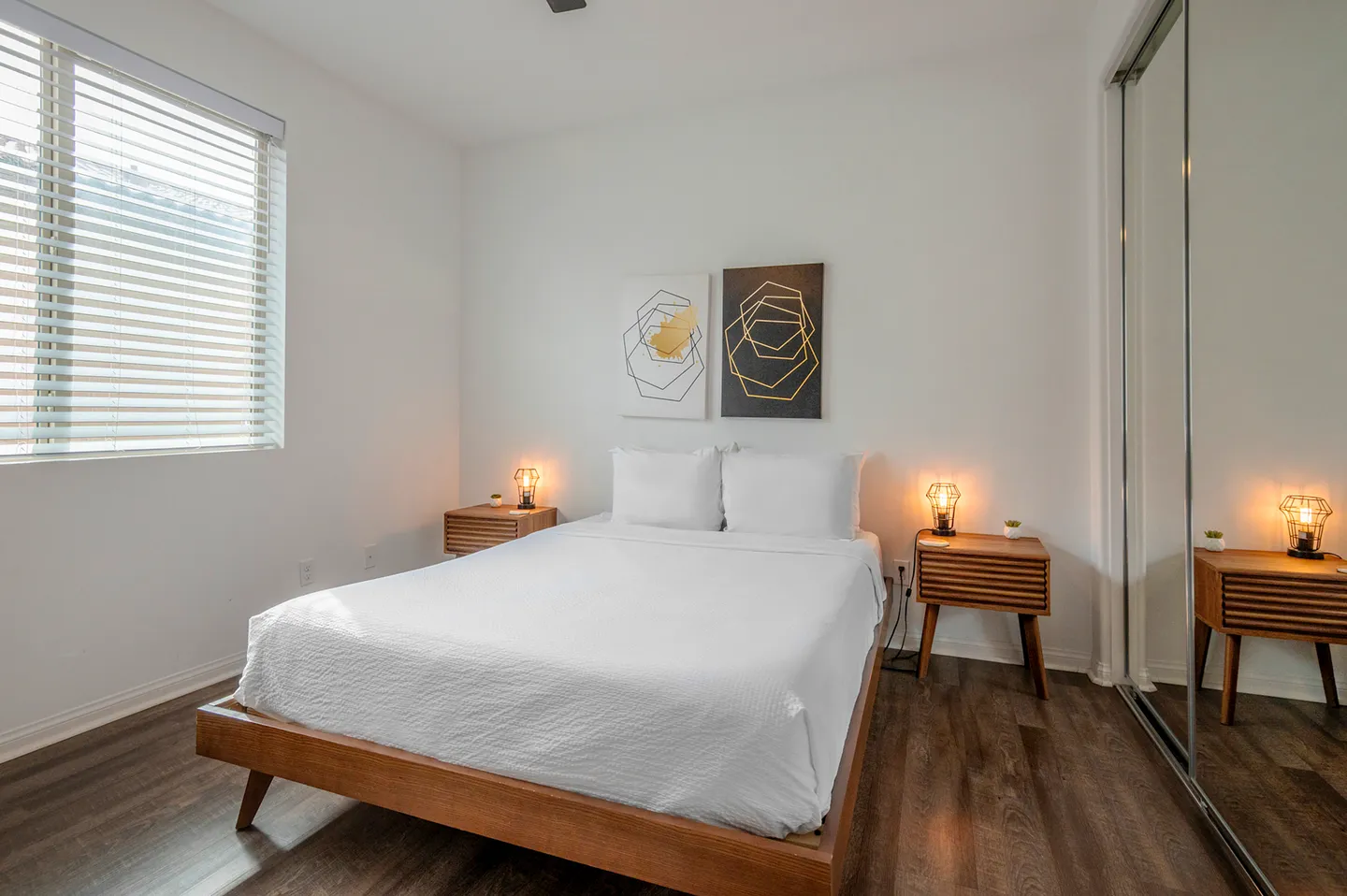 Bedroom with a wooden platform bed made with white linens, flanked by matching nightstands with glowing lamps, next to a mirrored closet and a window with blinds.