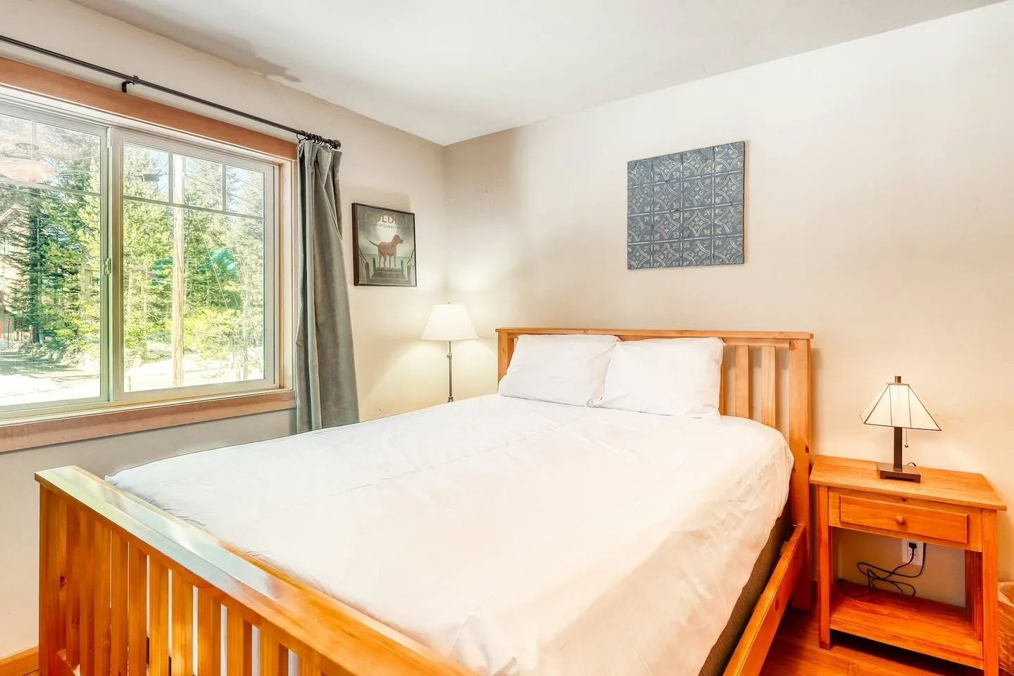 Bedroom with a wooden bed, white bedding, nightstand with lamp, and a large window looking out onto trees.