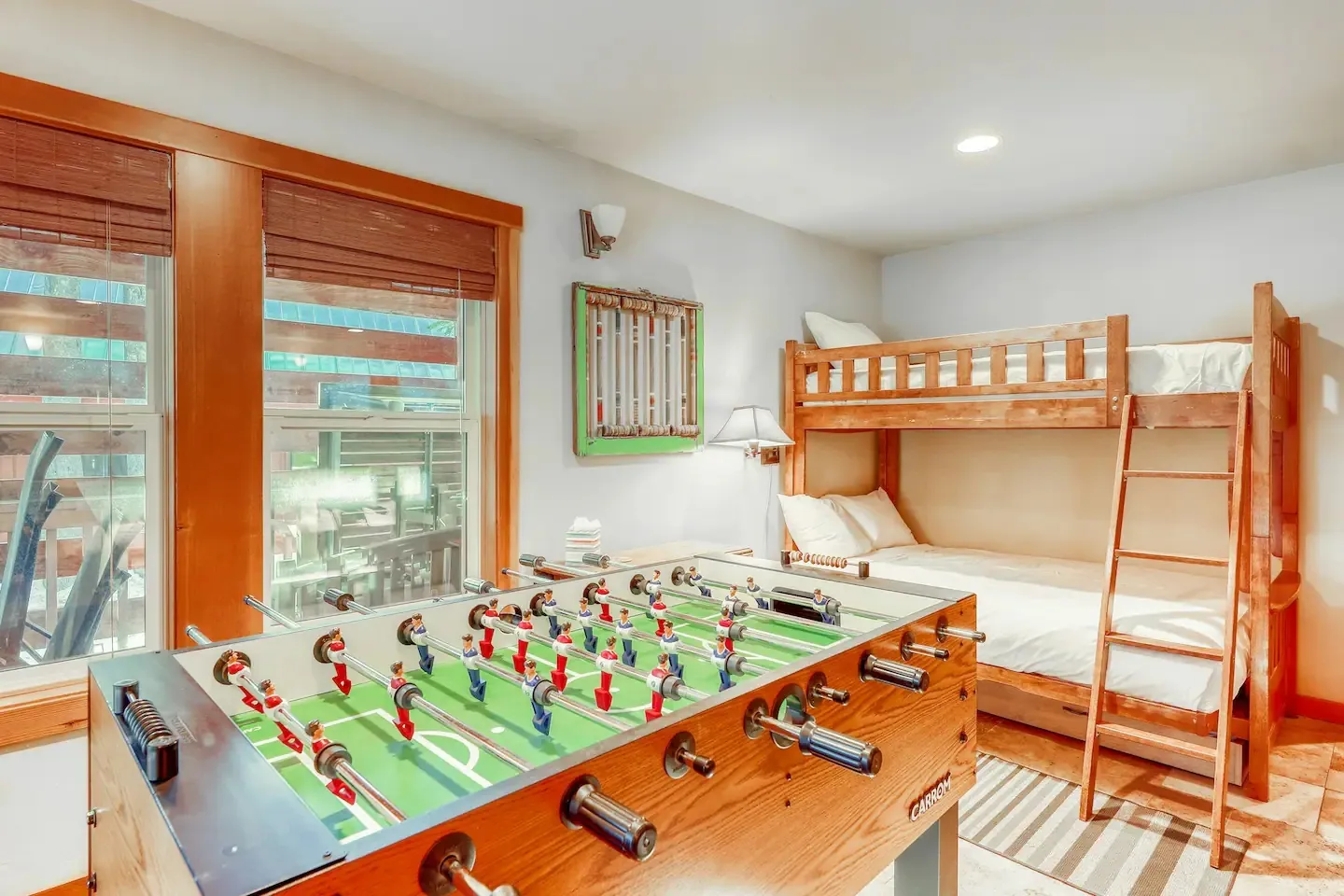 A foosball table positioned in a room with a wooden bunk bed and a window with blinds.