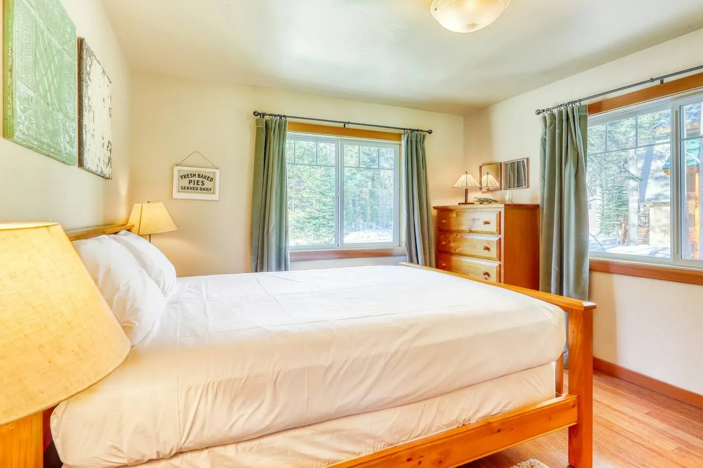 Bedroom with a wood-framed bed made with white linens, two windows framed by green curtains, a wooden dresser, and wood-look flooring.