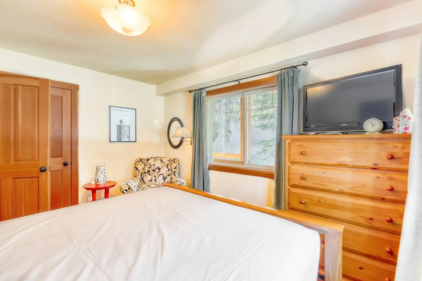 Bedroom with a wooden bed frame, a tall wooden dresser holding a TV, a patterned armchair, and a window with curtains.