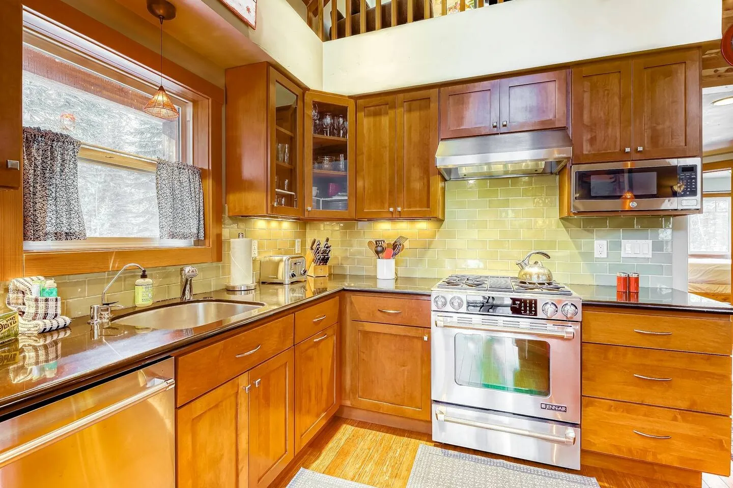 Kitchen corner showing wooden cabinetry, stainless steel appliances, a green tile backsplash, and a sink beneath a window.