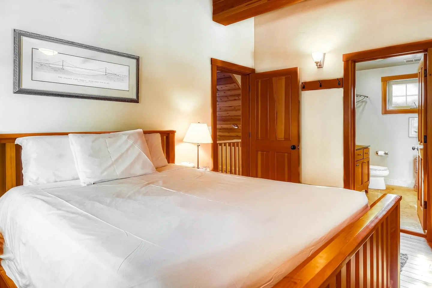 Bedroom with a wooden bed frame, framed bridge artwork above the headboard, and an open door leading to an en-suite bathroom.