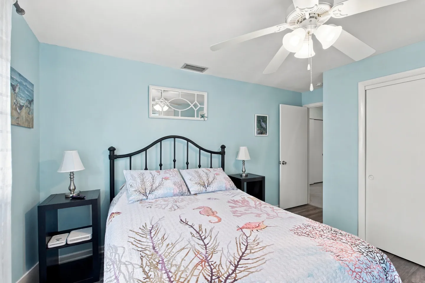 Bedroom with light blue walls, a black metal frame bed with coastal bedding, matching nightstands, and a ceiling fan.