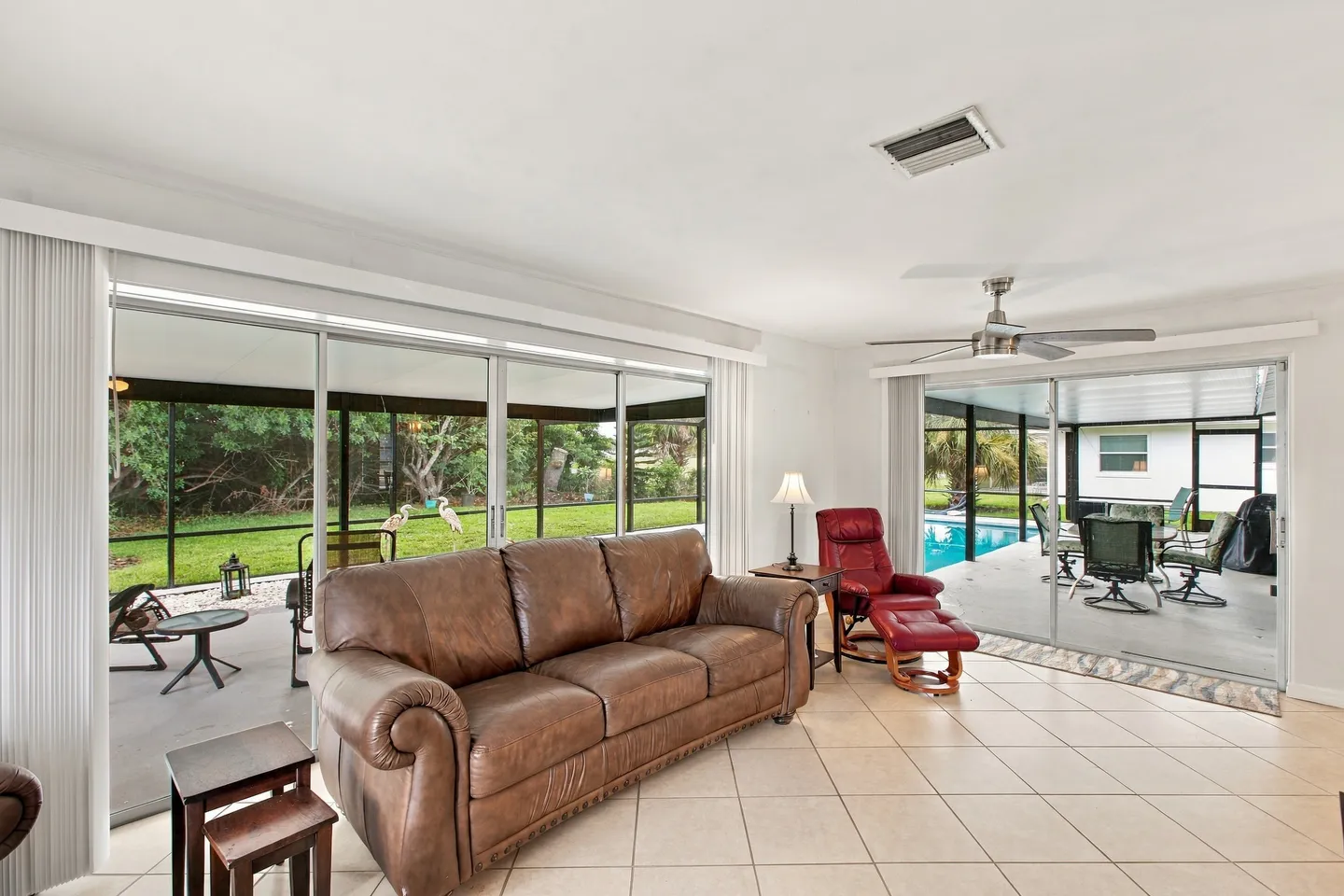 Living area with tiled floor, brown leather sofa, red recliner, and large sliding glass doors leading to a screened patio and pool enclosure.