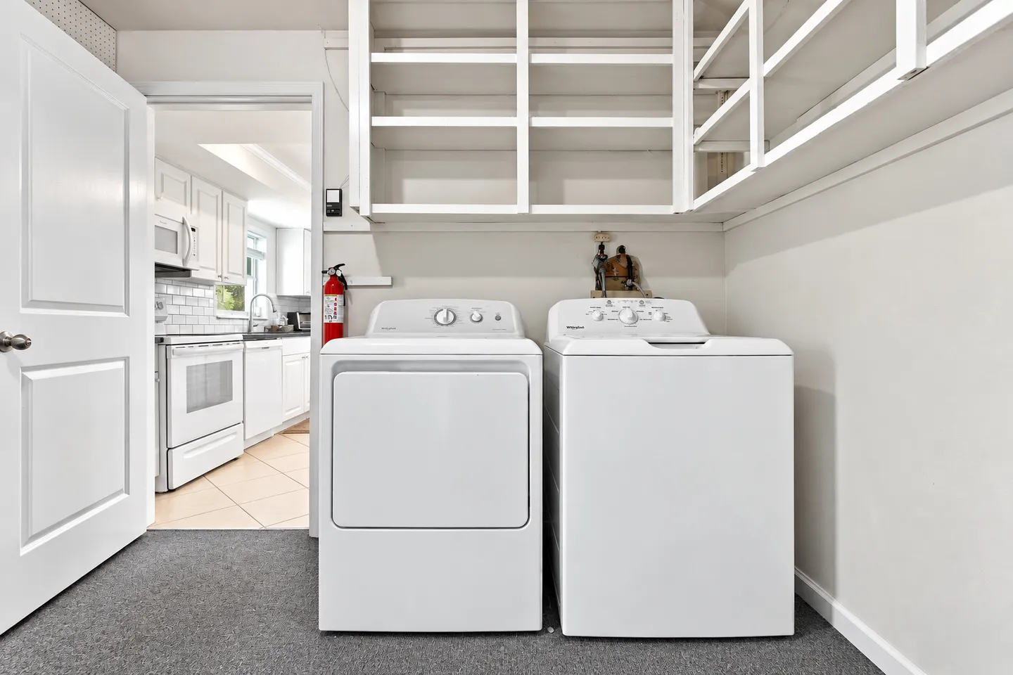 A laundry room with a white washer and dryer, open shelving above, and a doorway leading into the kitchen.