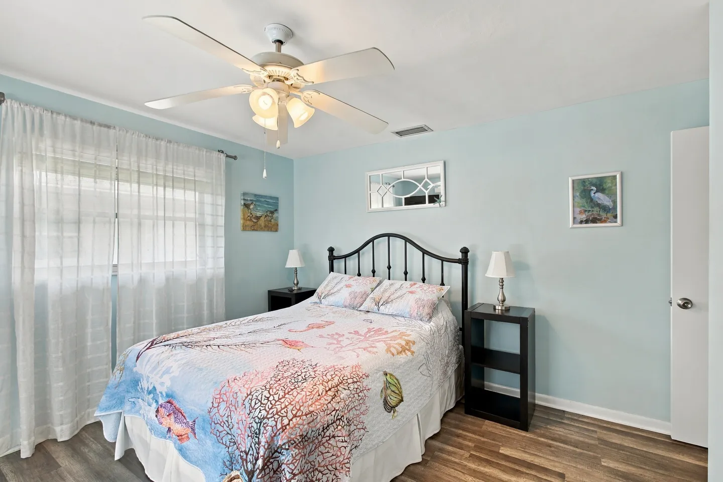 Bedroom with light blue walls, a bed with a coastal patterned quilt, a ceiling fan, and a window with sheer curtains.
