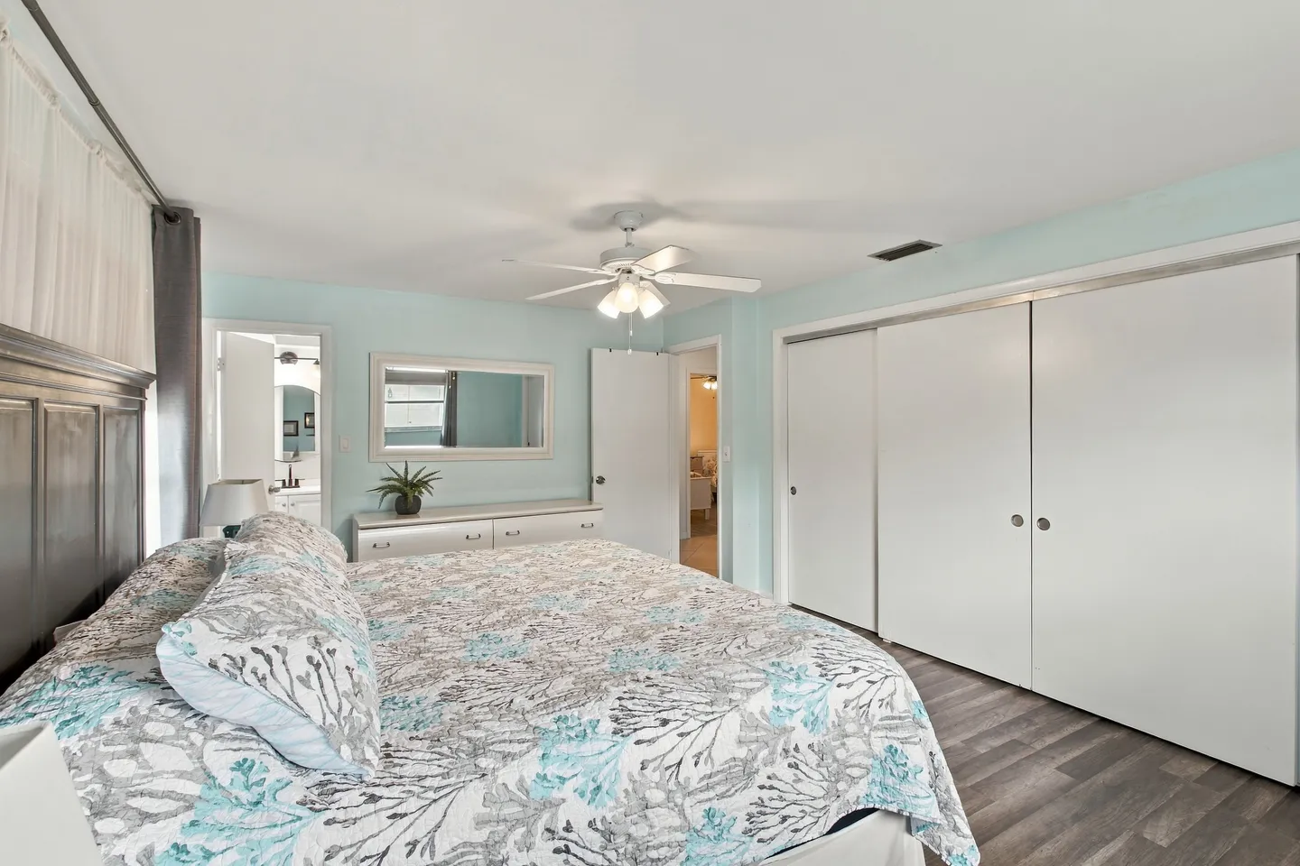 Bedroom with coastal-patterned bedspread, sliding closet doors, ceiling fan, and view into an en-suite bathroom.