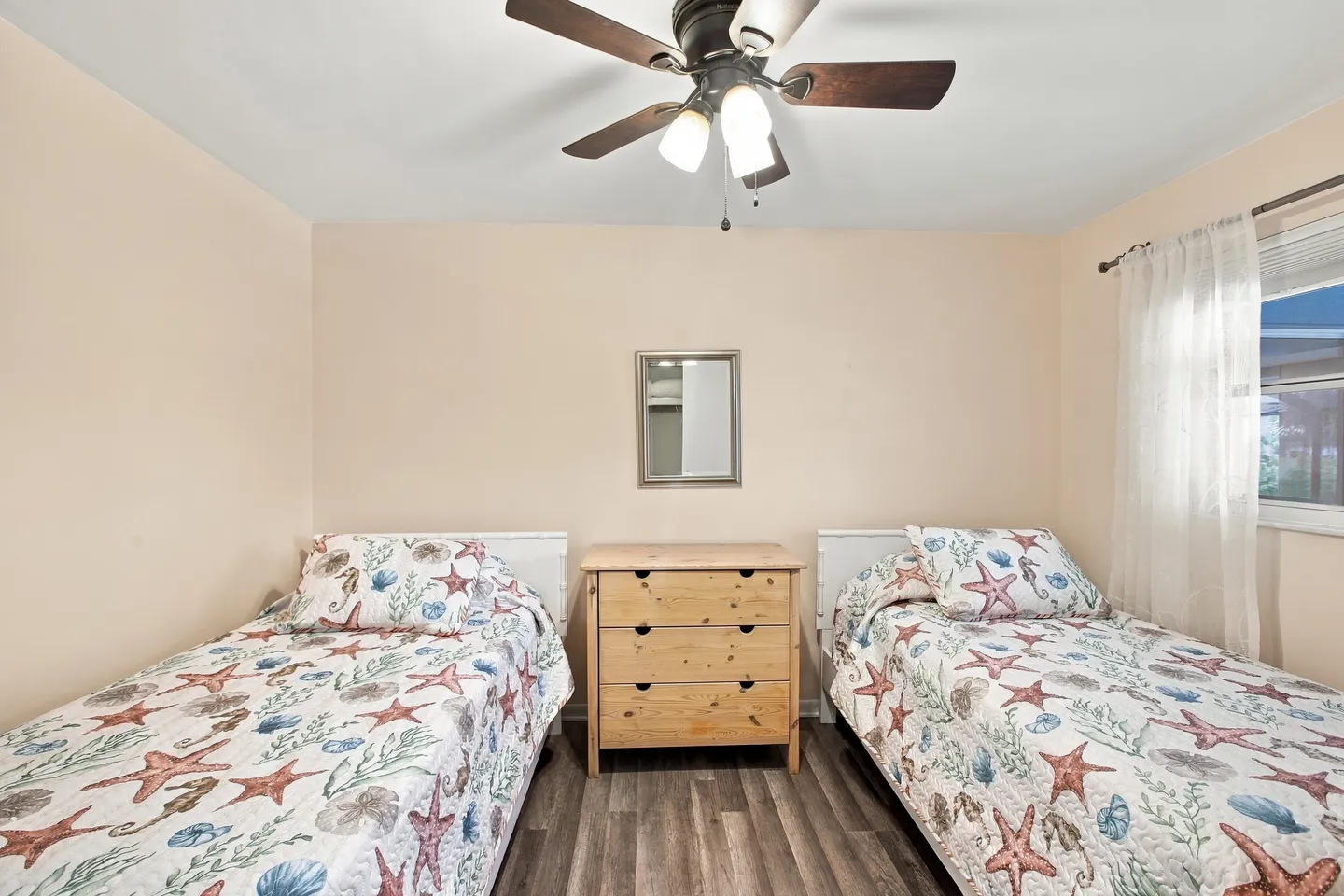 Bedroom with two twin beds dressed in coastal-themed quilts separated by a wooden dresser beneath a mirror and ceiling fan.