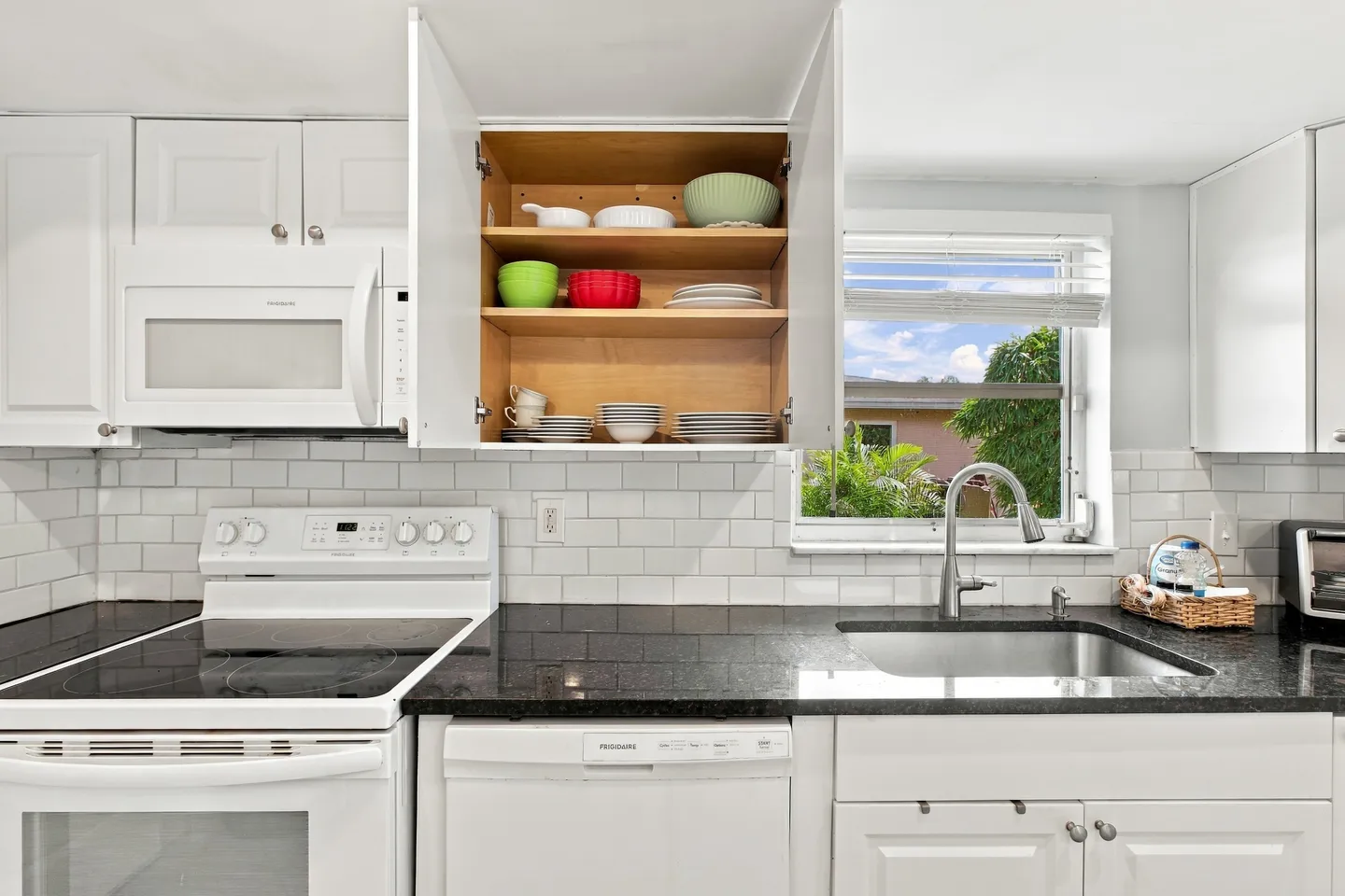 Kitchen counter with white cabinets, dark countertops, an open upper cabinet displaying bowls and plates, and a window above the sink.