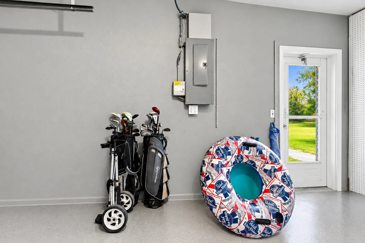 Garage interior showing golf bags on a push cart, an inflatable pool tube, an electrical panel, and an exterior door leading to a yard.
