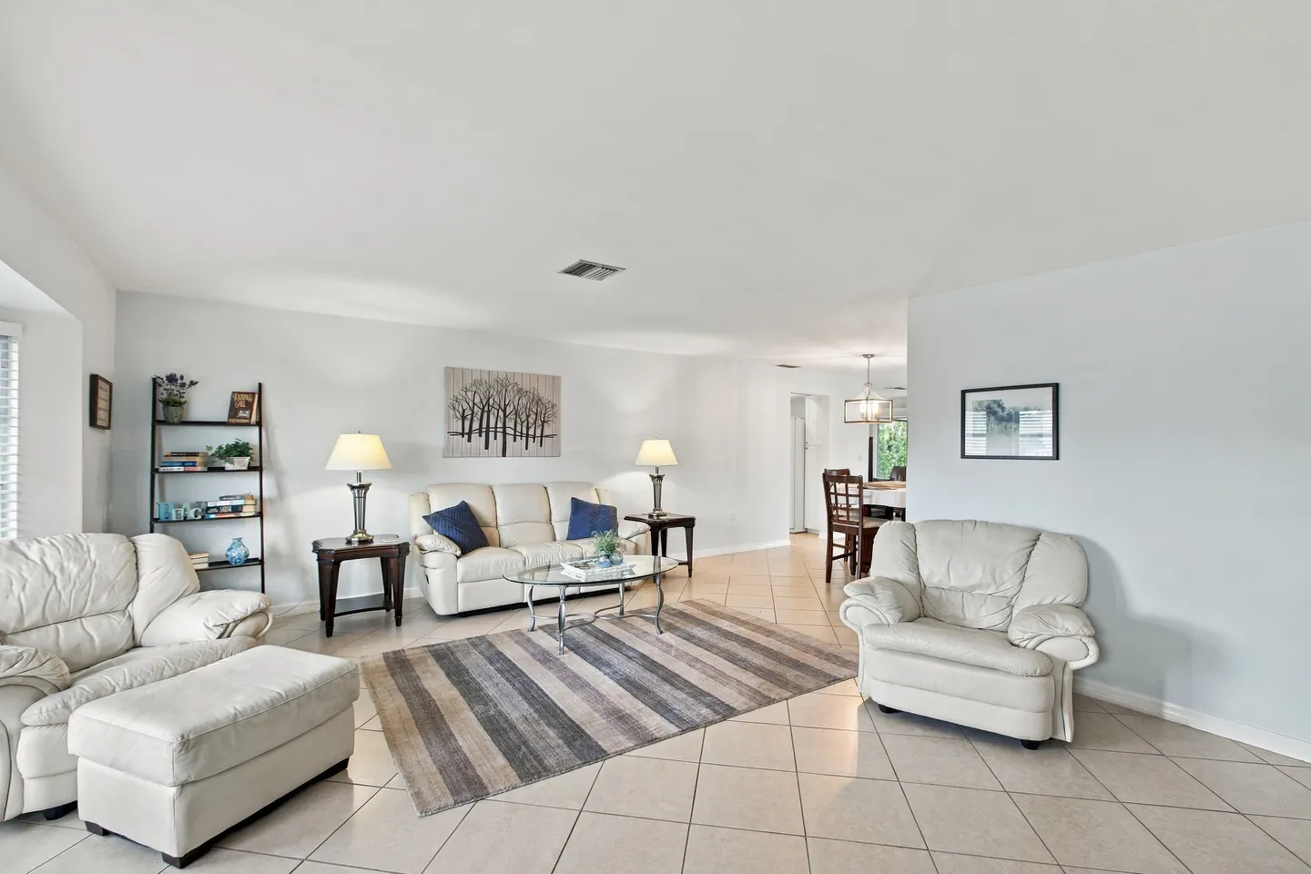 Living room with light-colored tile flooring, cream leather seating, a striped area rug, and an adjacent dining area.