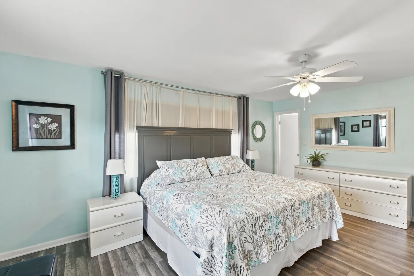 Bedroom with light blue walls, a bed with a dark gray headboard, white dressers, and a ceiling fan.