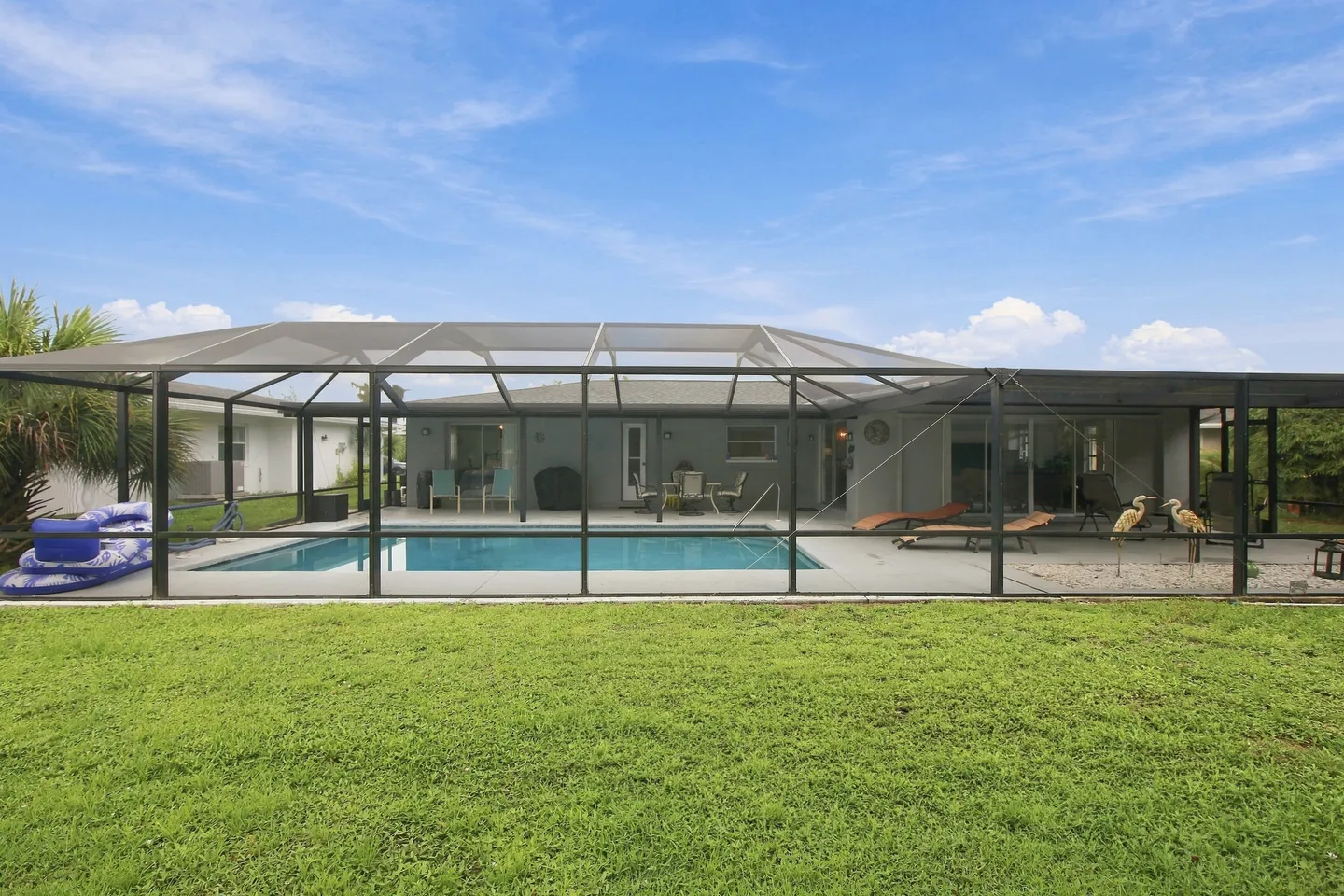 Exterior view of a home's backyard with a grass lawn in the foreground and a large screened pool enclosure with a swimming pool and patio furniture.
