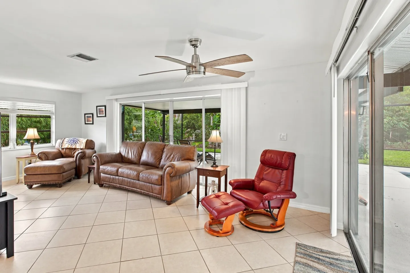 Living room with tiled floor, brown leather sofa, armchair with ottoman, red leather recliner, and sliding glass doors leading outside.