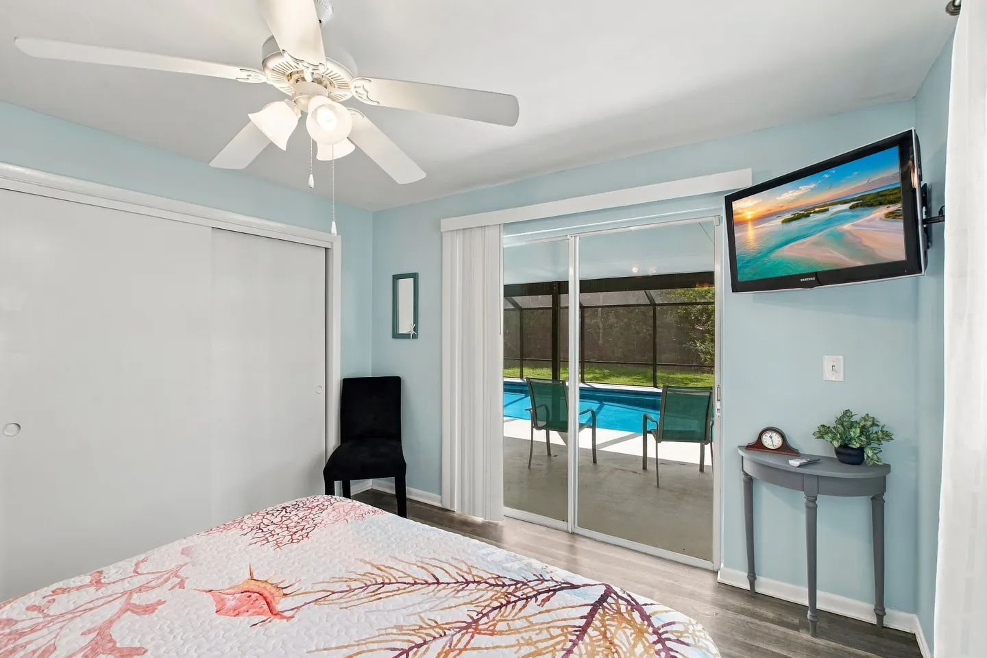 Bedroom with light blue walls, ceiling fan, and sliding glass doors opening to a covered patio and screened-in swimming pool.
