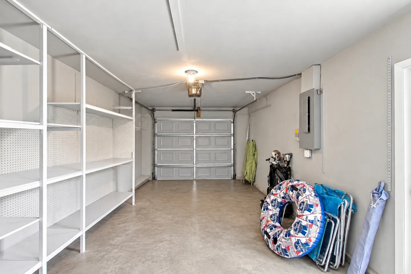 A clean, bright single-car garage with white shelving units on the left wall, concrete flooring, and beach gear stacked on the right.