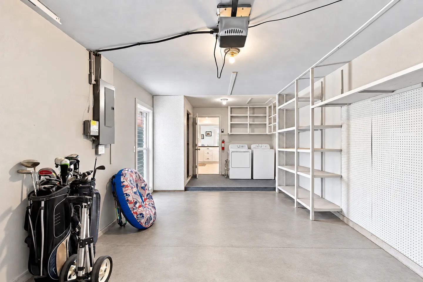 Interior view of an organized garage featuring concrete flooring, tall white shelving units, a washer and dryer area, and golf equipment.