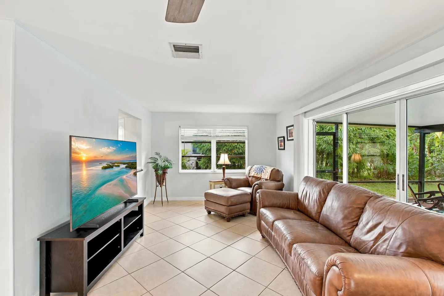 Living room with tiled floor, brown leather couch, armchair with ottoman, flat-screen television on a dark media console, and sliding glass doors.