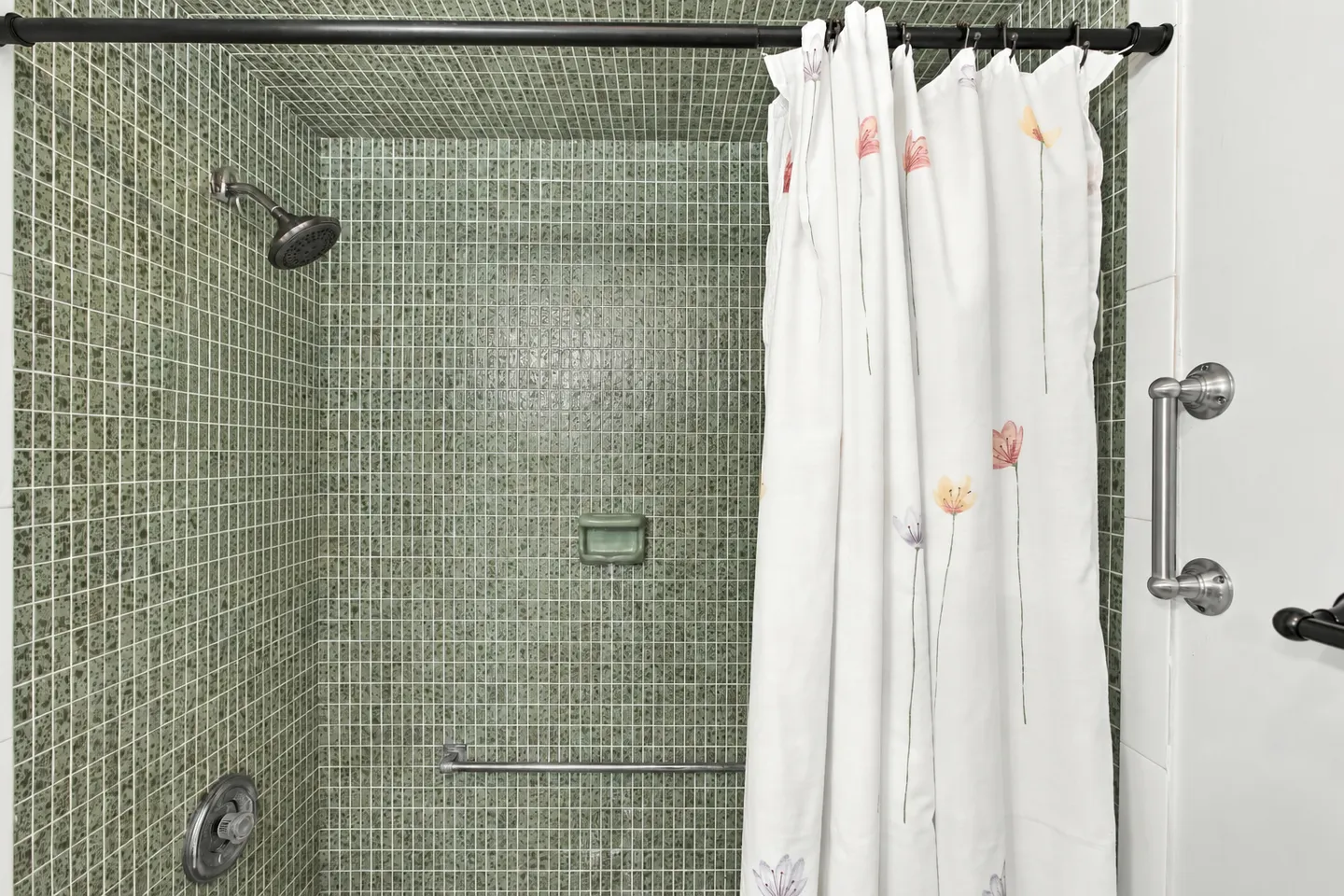 A walk-in shower lined with small green mosaic tiles, featuring a dark showerhead, towel bar, and a partially drawn floral shower curtain.