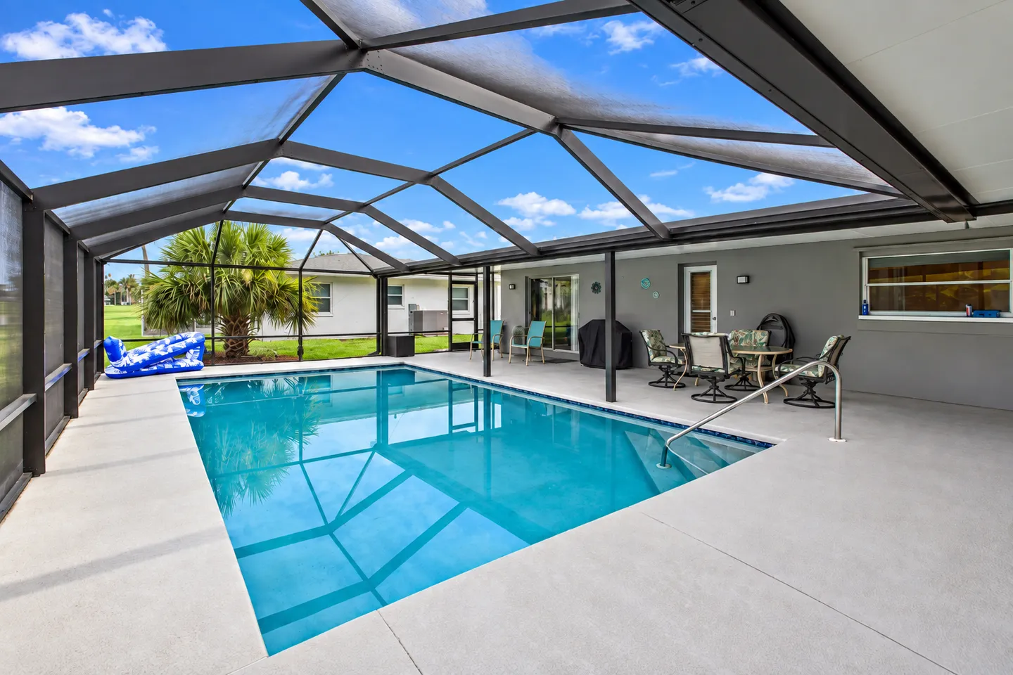 Screened-in outdoor swimming pool and patio with outdoor dining furniture against a house wall under a blue sky.