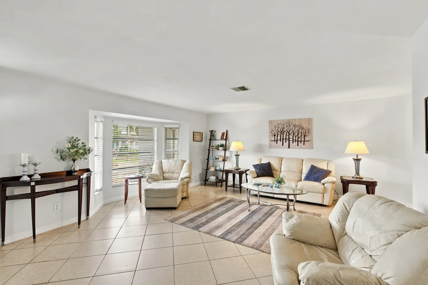 Bright living room with tiled flooring, neutral-toned leather seating, a bay window with blinds, and light-colored walls.