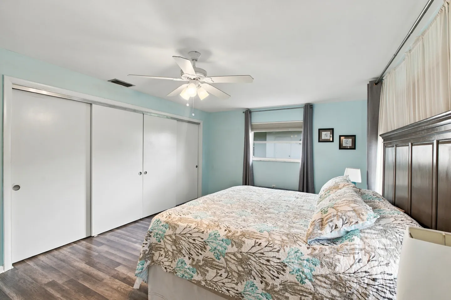 Bedroom with light blue walls, a bed with a floral quilt, wood-style flooring, a ceiling fan, and sliding closet doors.