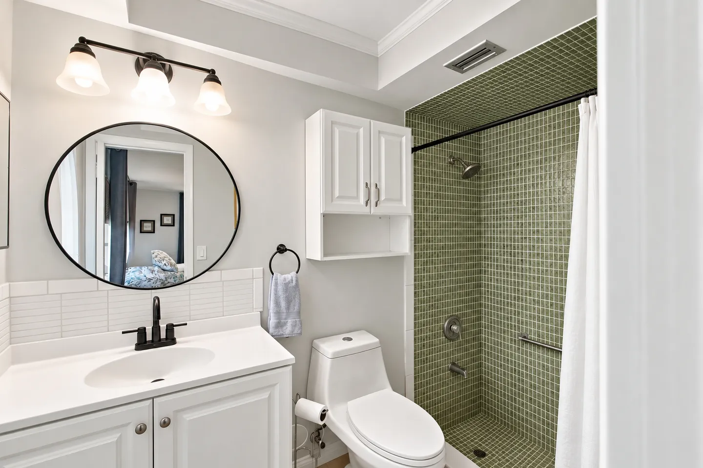 Bathroom with white vanity, round mirror, toilet, overhead cabinet, and a walk-in shower with green mosaic tile.
