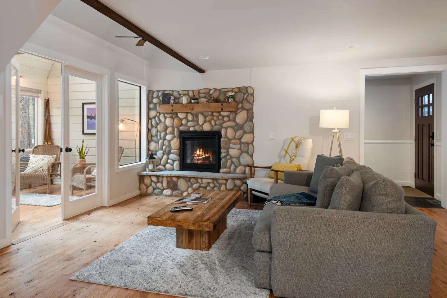 Living room featuring a corner stone fireplace with an active fire, wood coffee table, gray sofa, and glass French doors leading to an adjacent sitting area.