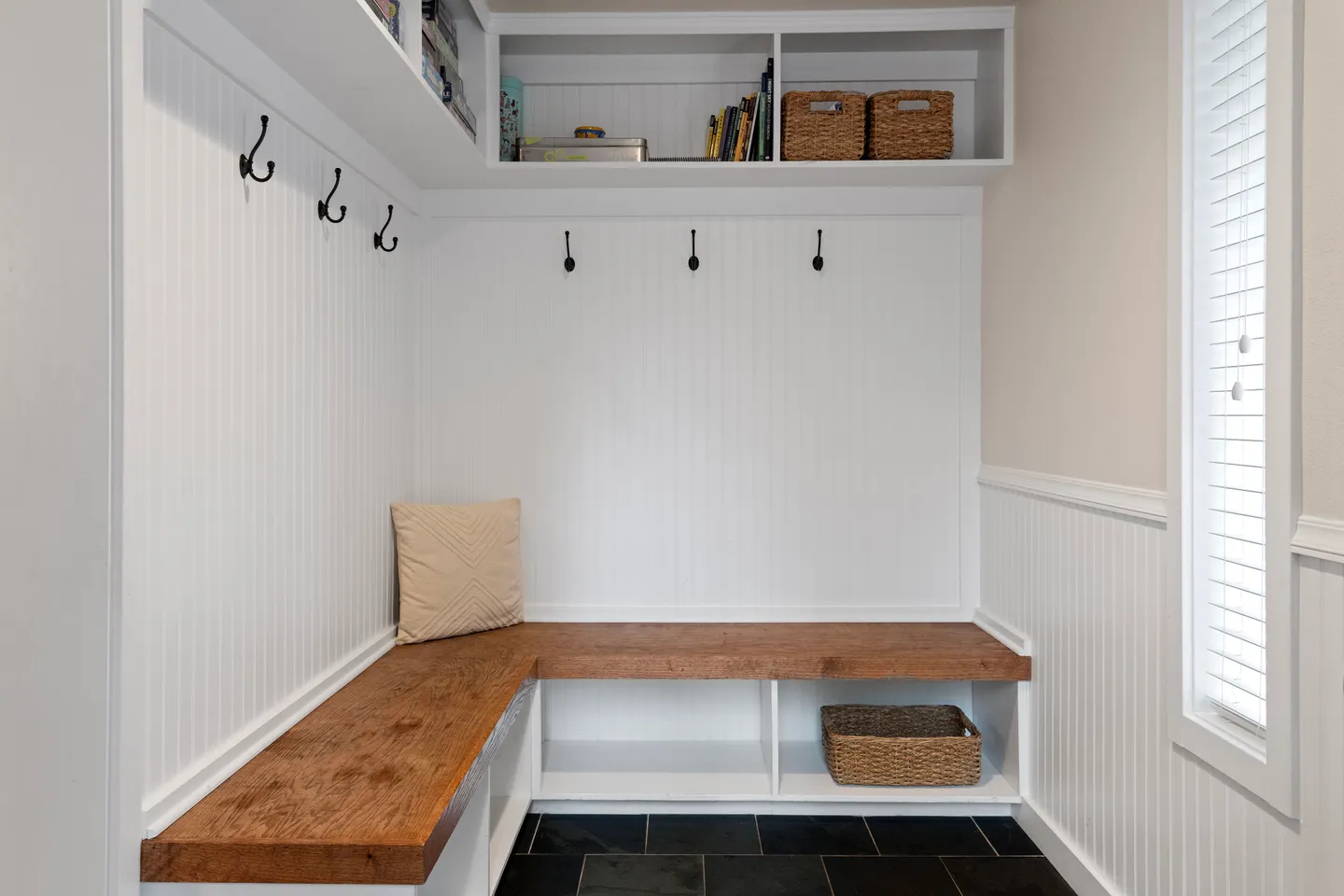Built-in mudroom area with an L-shaped wooden bench, wall hooks, upper cubbies with storage baskets, and dark tile flooring.