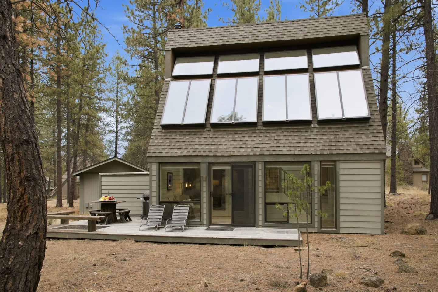 Exterior view of a multi-story cabin with a steep roof featuring skylight panels, a ground-level deck, and surrounding pine trees.