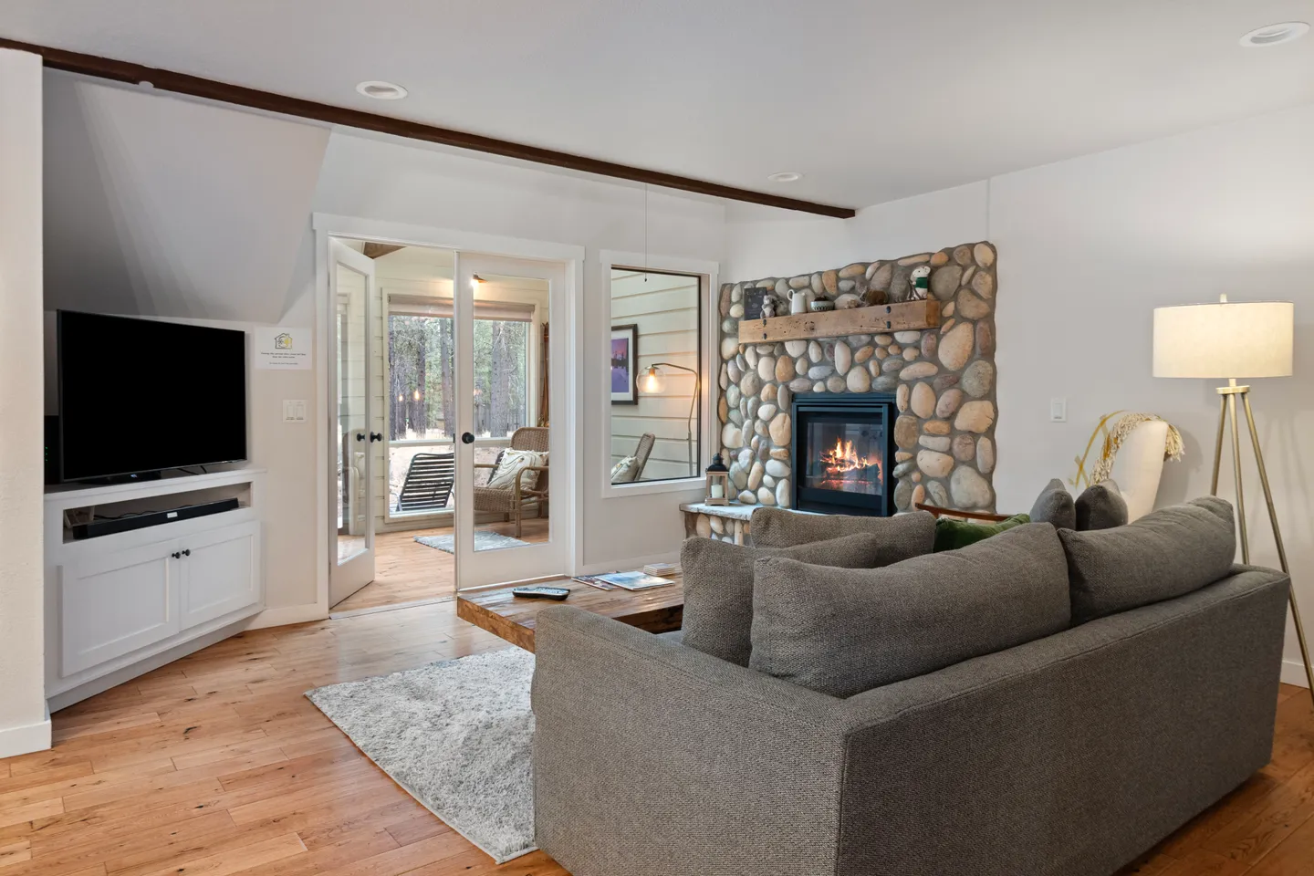 Living room with a stone-surround fireplace, a gray sofa, a television on a white stand, and French doors leading to an enclosed porch.