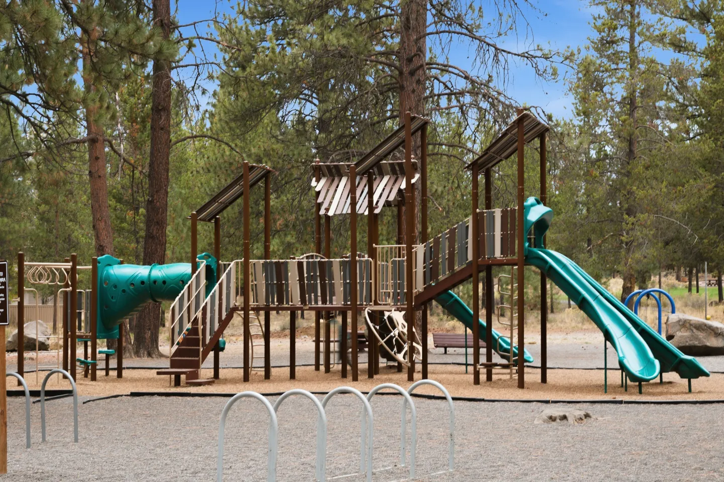 Outdoor children's playground structure with green slides, climbing elements, and bike racks set amidst pine trees.
