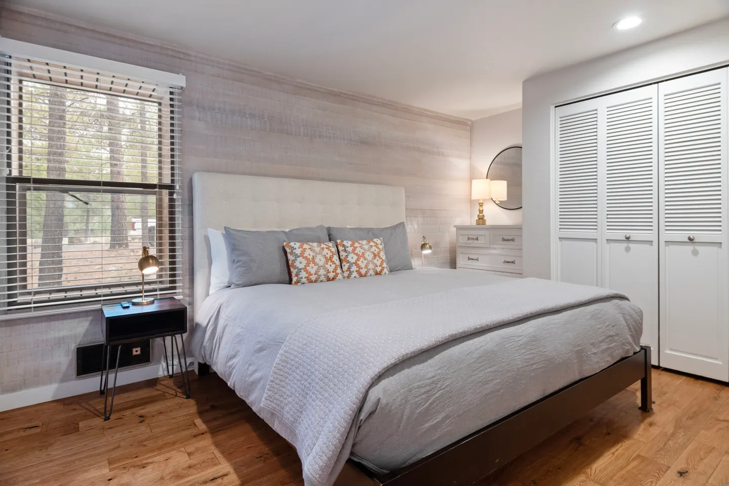 Bedroom with an upholstered headboard, grey bedding, wood-look accent wall, window with blinds, and white louvered closet doors.