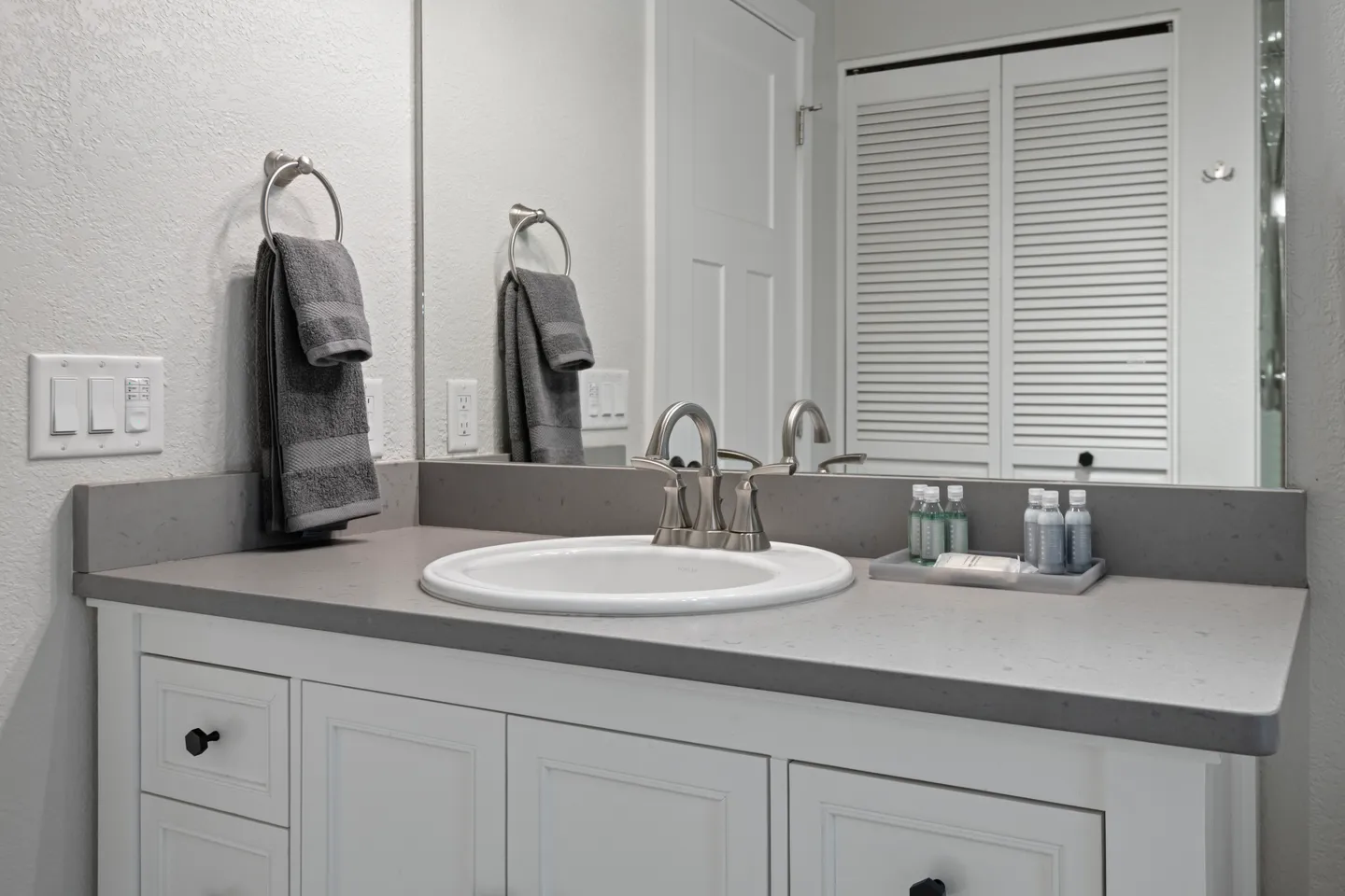 Bathroom vanity with a gray countertop, white sink, brushed metal faucet, large mirror, and white cabinetry.