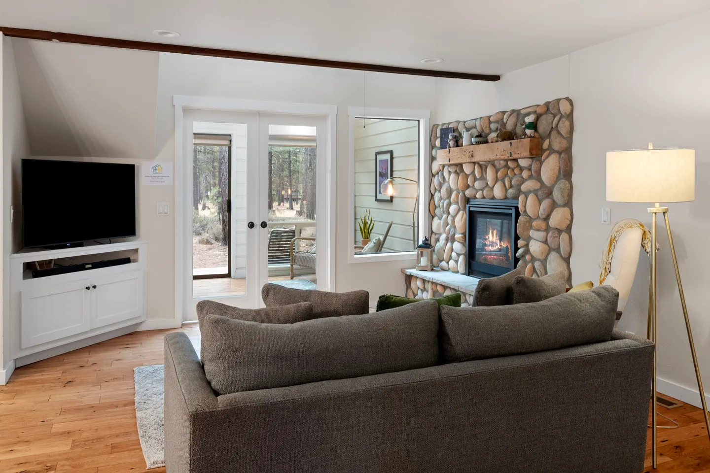 Living room featuring a grey sofa, river rock fireplace, television on a white console, and glass doors leading outside.