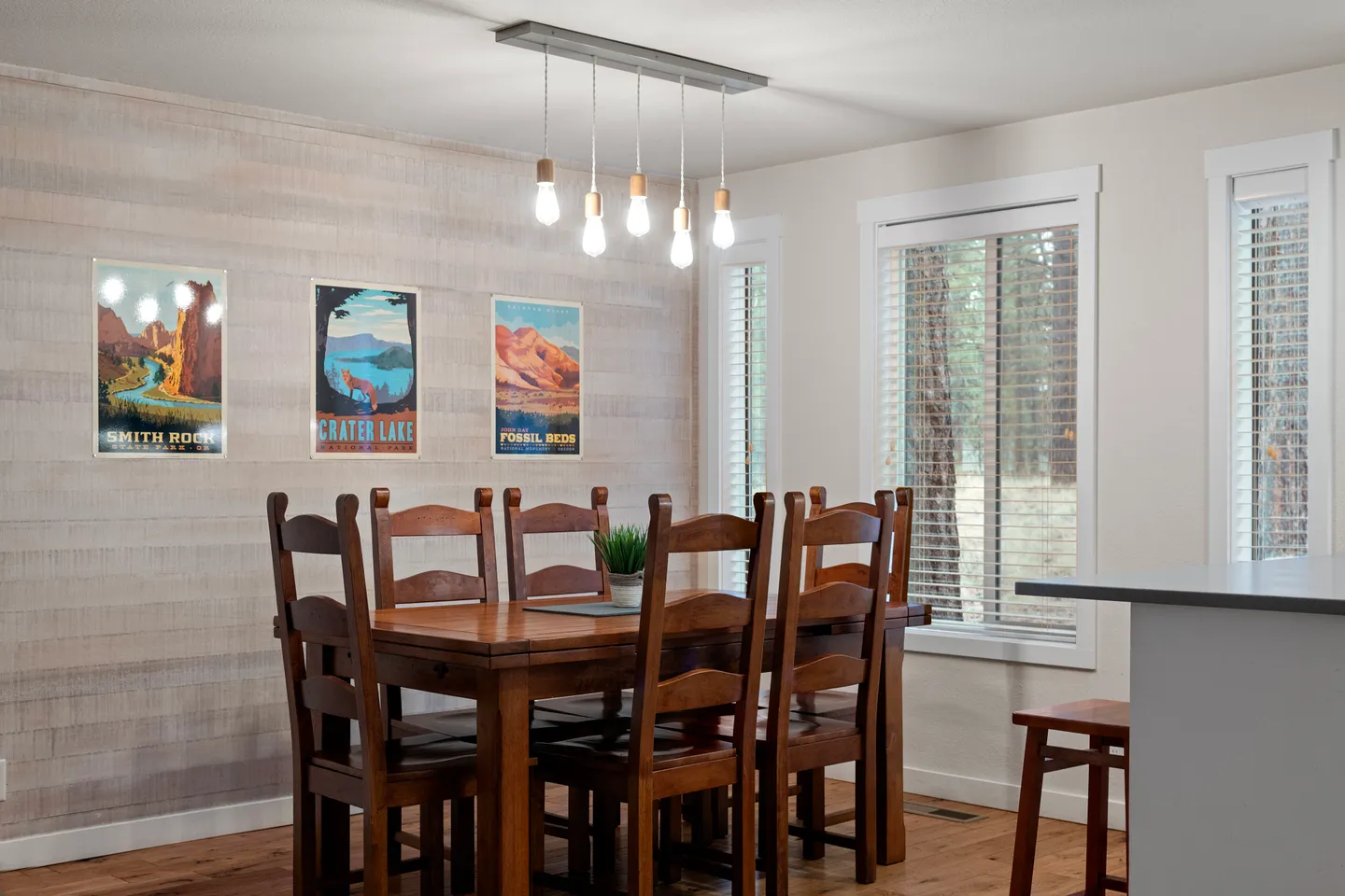 Dining area with a wooden table and chairs beneath a modern hanging bulb light fixture, set against an accent wall with travel posters.