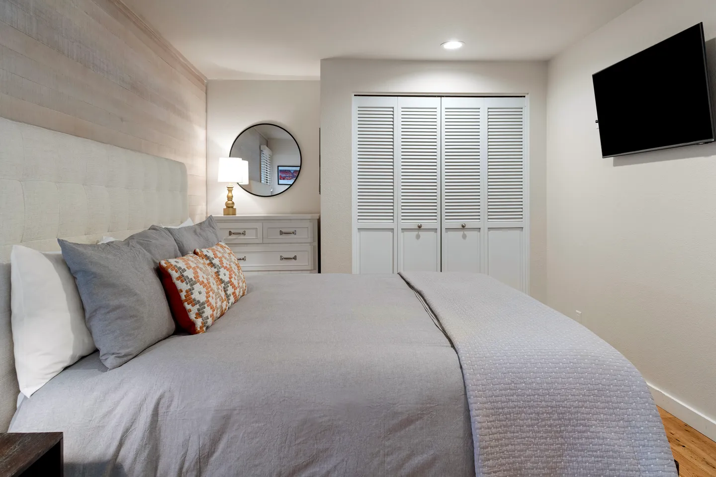 Bedroom featuring a bed with gray bedding, a wall-mounted TV, white louvered closet doors, and a dresser with a round mirror.