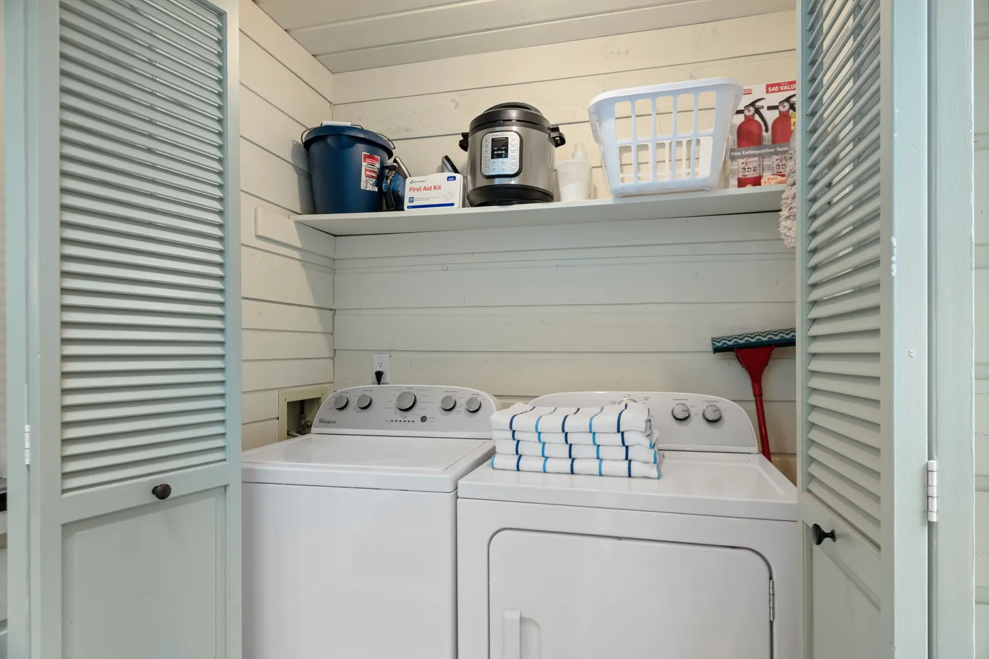 A laundry closet behind louvered doors with a white washer and dryer, striped towels, and an upper shelf with supplies.