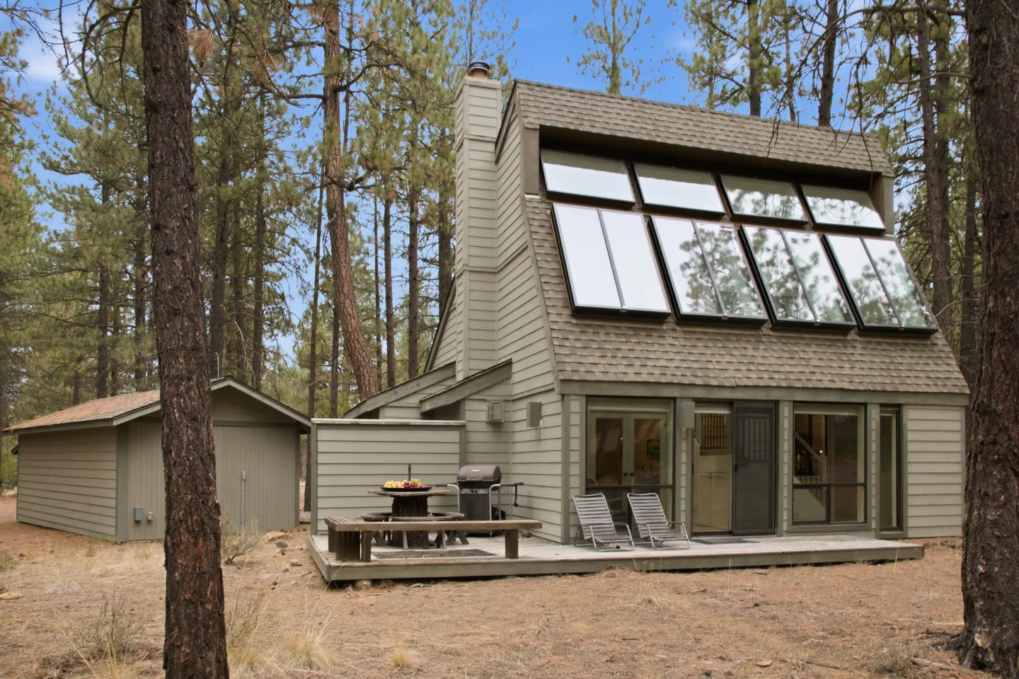 Exterior view of a cabin with a sloped roof featuring large angled windows, set on a wooden deck in a pine forest.