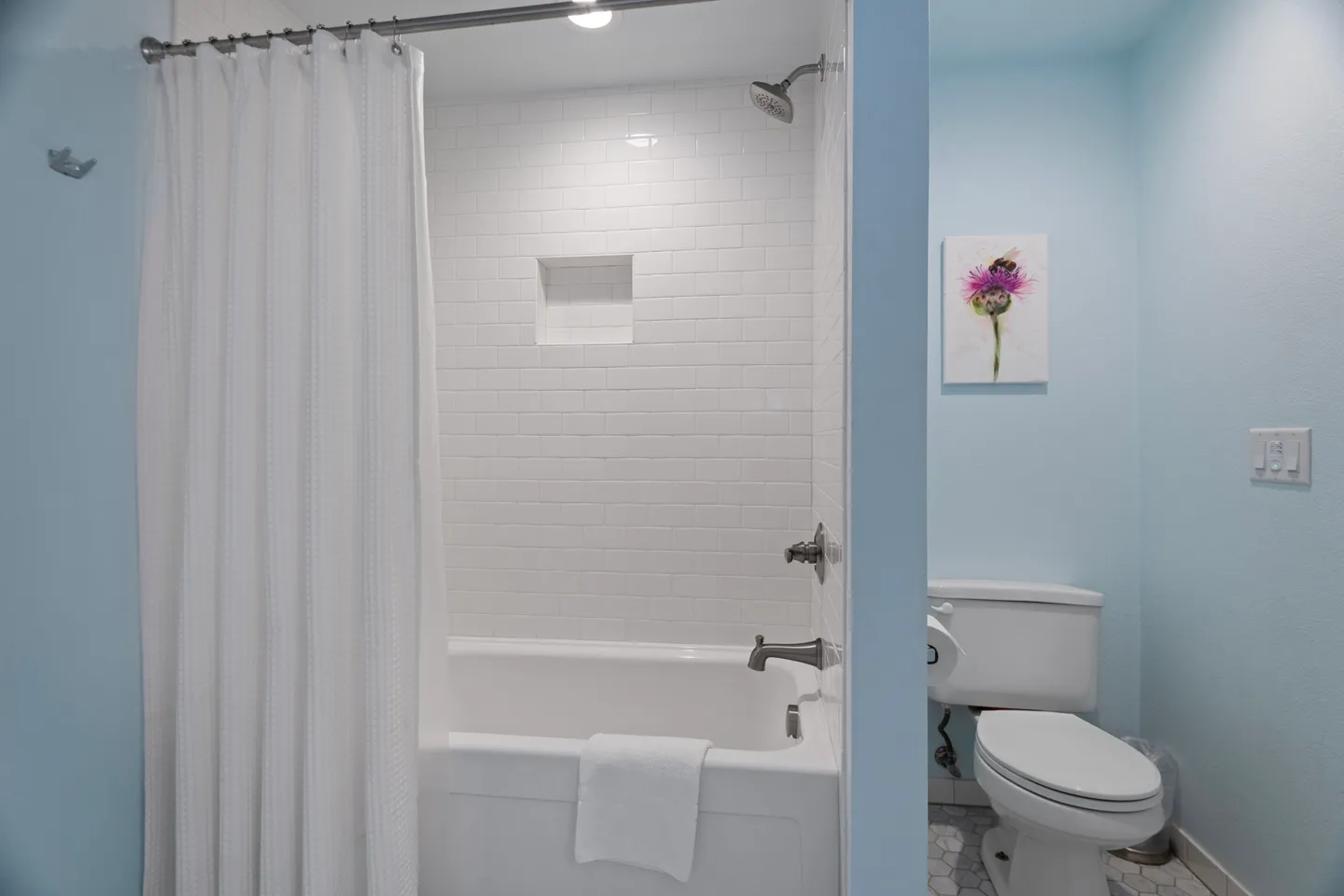Bathroom showing a white bathtub shower combination lined with white subway tile alongside a toilet against light blue walls.