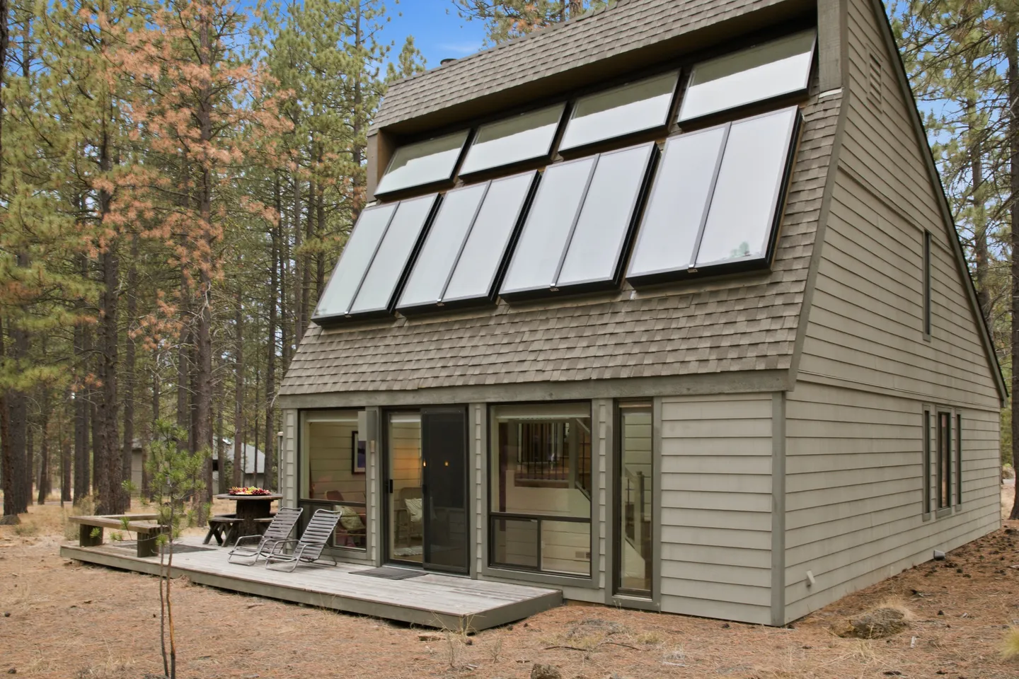 Exterior view of an A-frame-style house with rooftop solar panels, a wooden deck with lounge chairs, and surrounding pine trees.