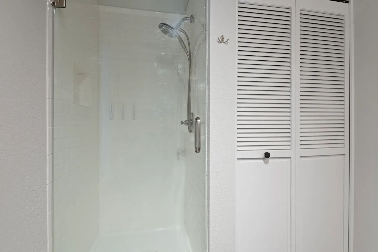 A walk-in shower with white subway tile, a chrome showerhead with handheld attachment, and a glass door next to white louvered closet doors.