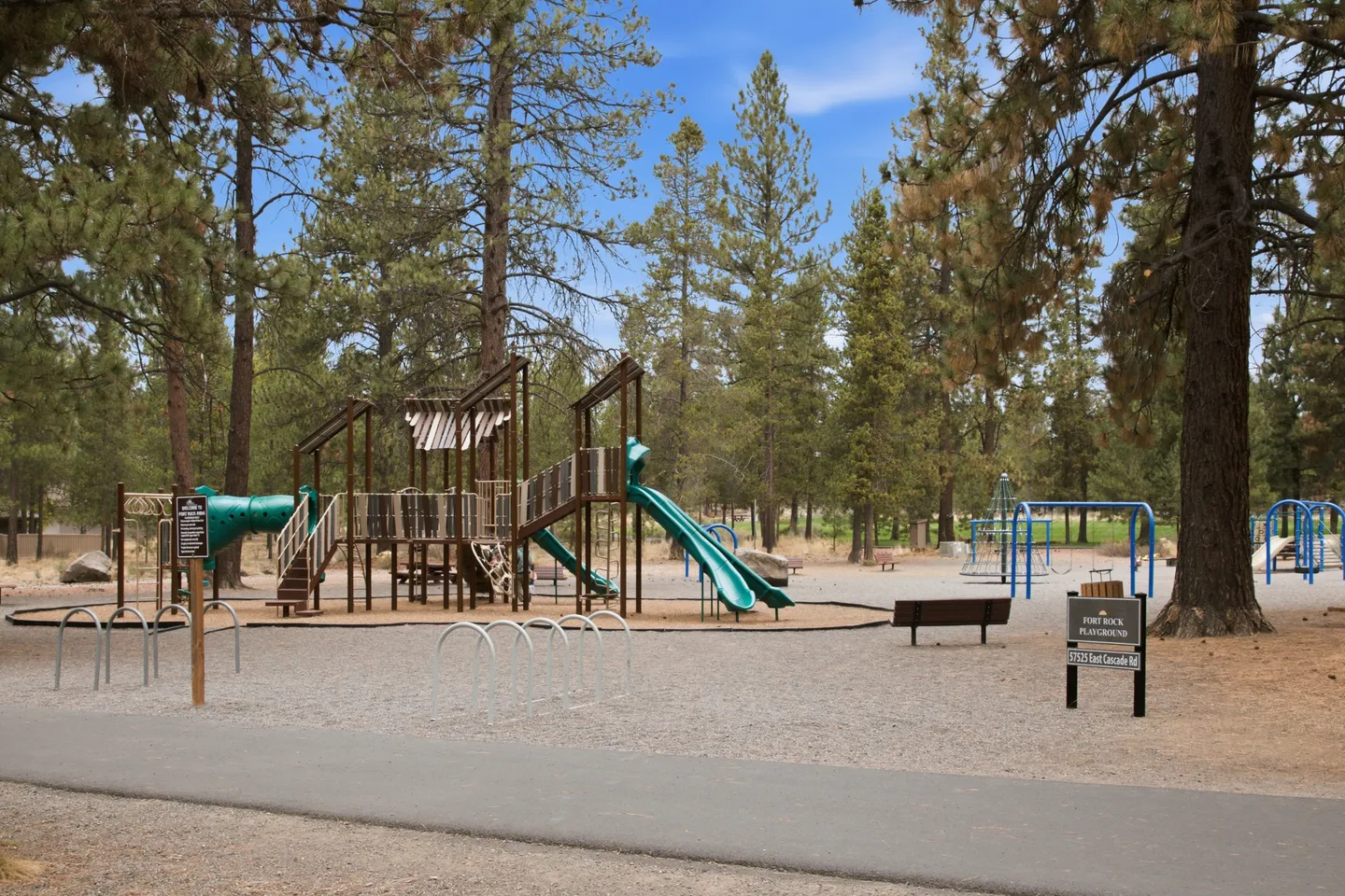 Outdoor playground surrounded by pine trees, featuring play structures with green slides, swings, and bike racks.