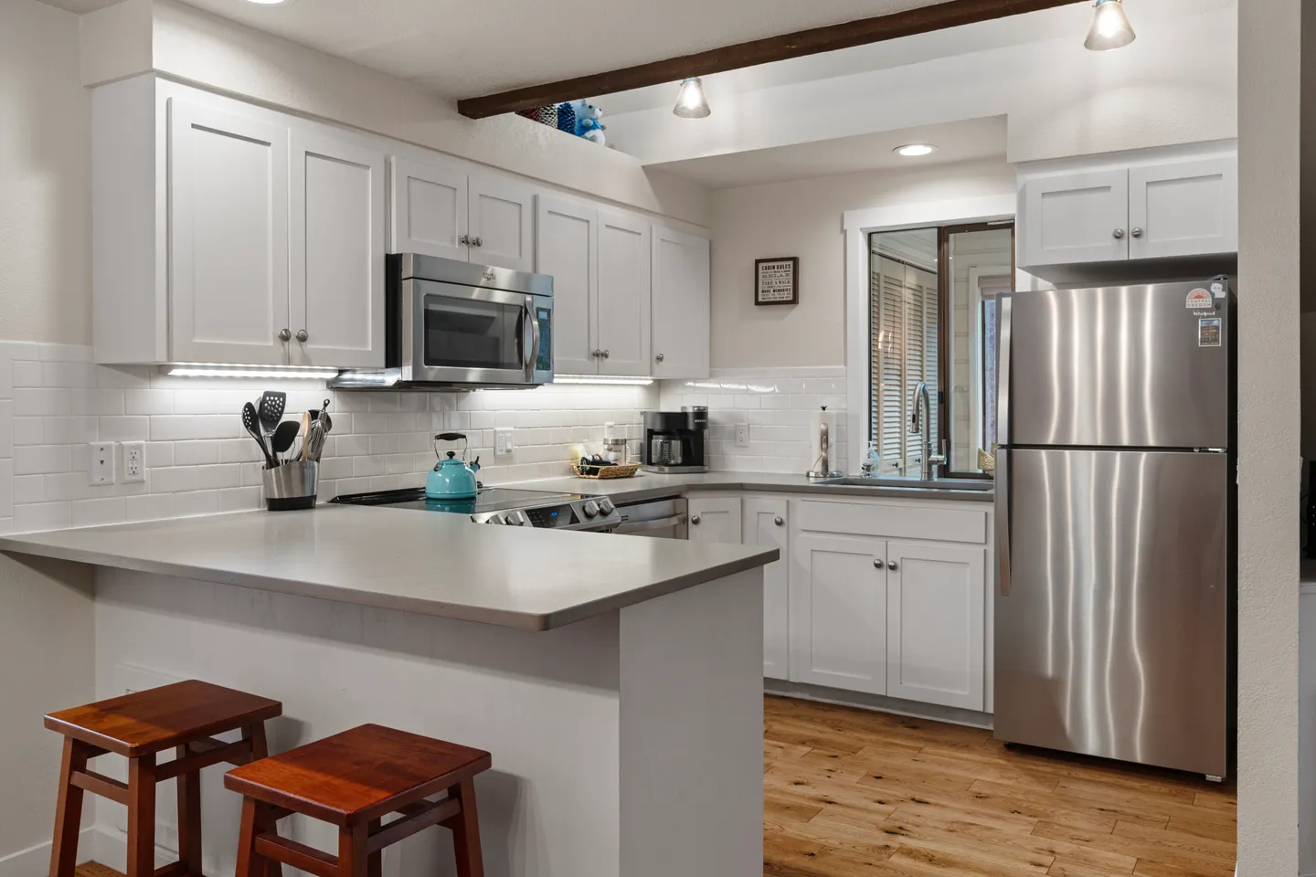 Kitchen with white shaker cabinets, stainless steel appliances, a breakfast bar with two wooden stools, and light wood flooring.