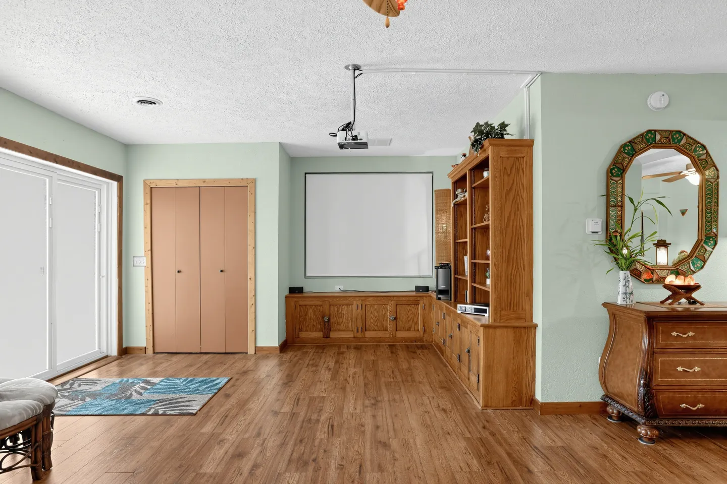 Living area featuring wood-look flooring, light green walls, a ceiling-mounted projector, a projection screen, and built-in wooden cabinets.