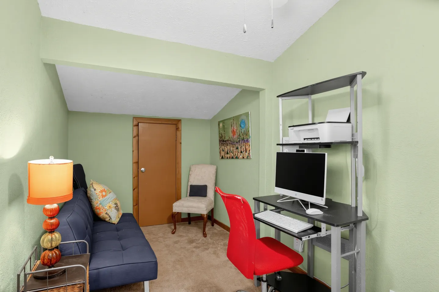 A small room or home office with light green walls, a blue futon sofa, a desk with a computer, and a red office chair.
