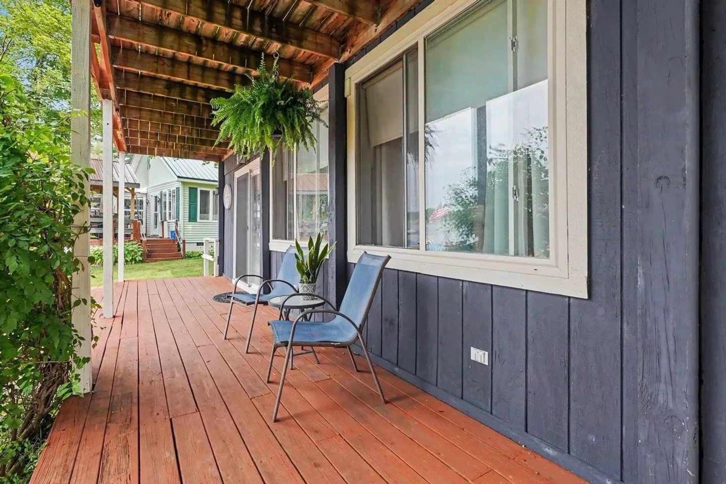 A covered wooden deck running alongside a dark blue exterior wall with sliding glass windows, outdoor chairs, and a hanging fern.