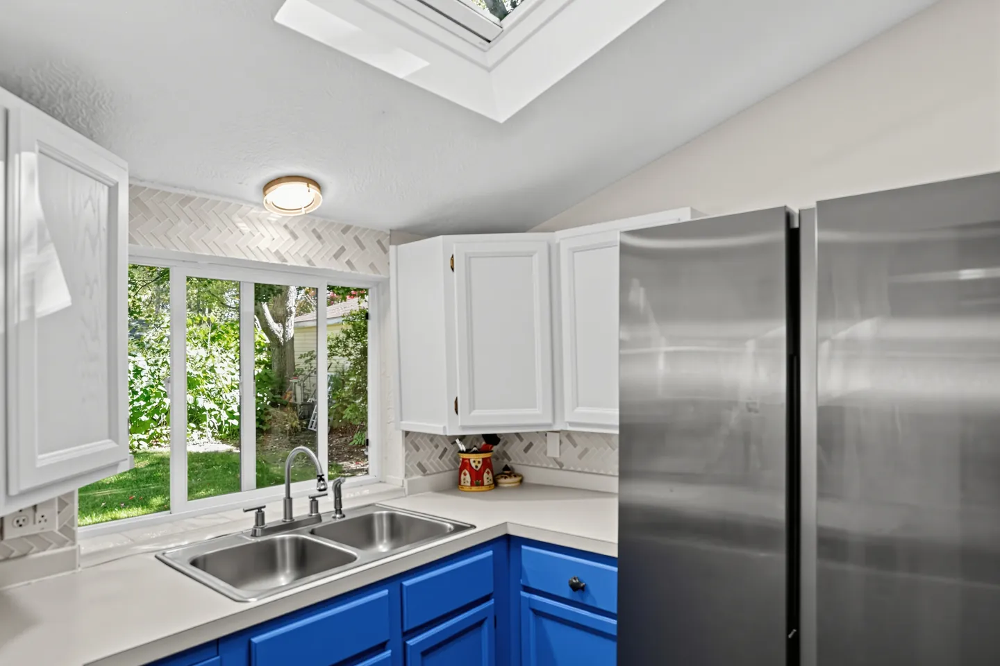 Kitchen featuring bright blue lower cabinets, white upper cabinets, a double stainless steel sink below a wide window, and a skylight.
